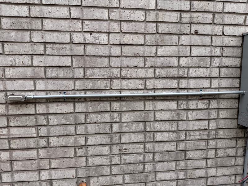 New galvanized steel conduit strapped across a brick wall from a service enclosure to a pulling elbow, with new conductors not yet landed at the enclosure