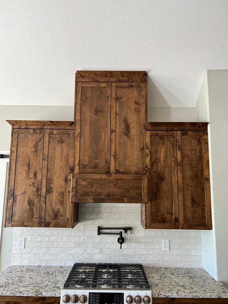 Stained alder range hood surround flanked by matching wall cabinets over a white brick-look backsplash and pot filler