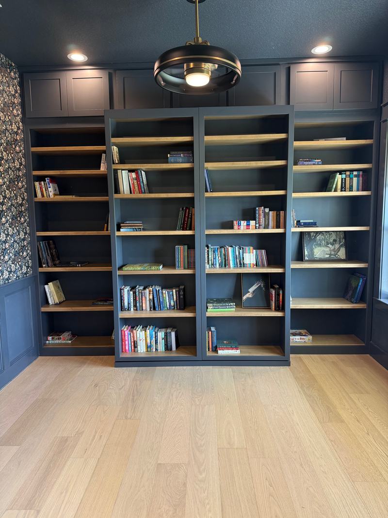 Navy built-in library wall with sliding center bookcases, stained wood shelves, shaker upper cabinets and a wide-plank oak floor