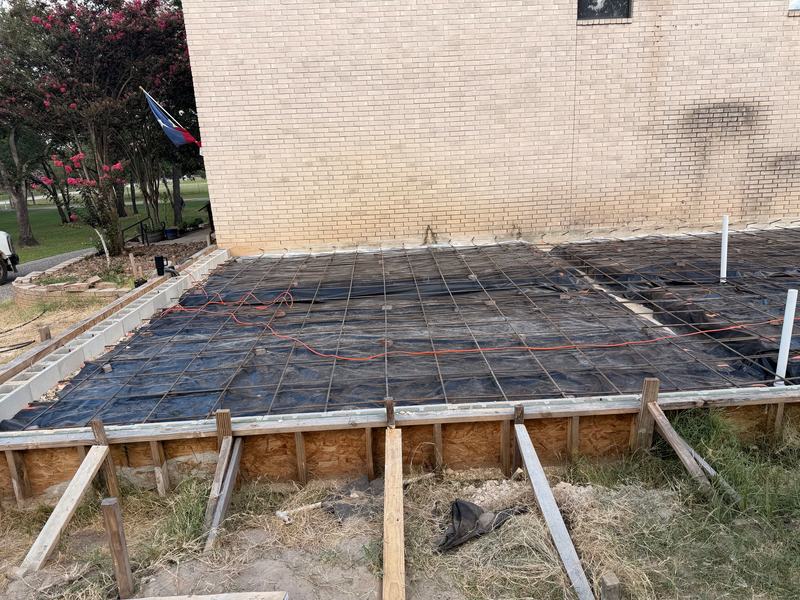 Rebar grid over a vapor barrier inside wood slab forms beside a brick wall, ready for the pour
