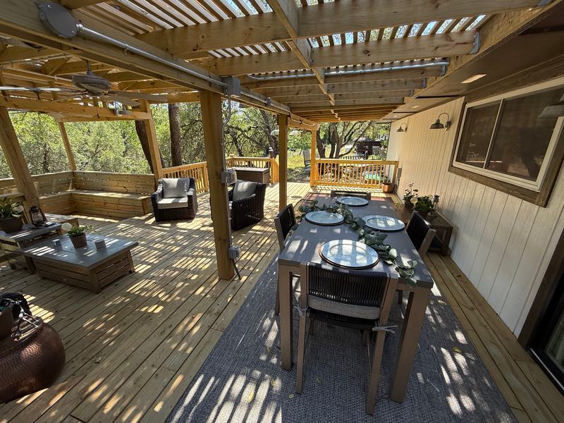 Wood deck under a slatted pergola with a dining table, lounge seating, ceiling fans and lights