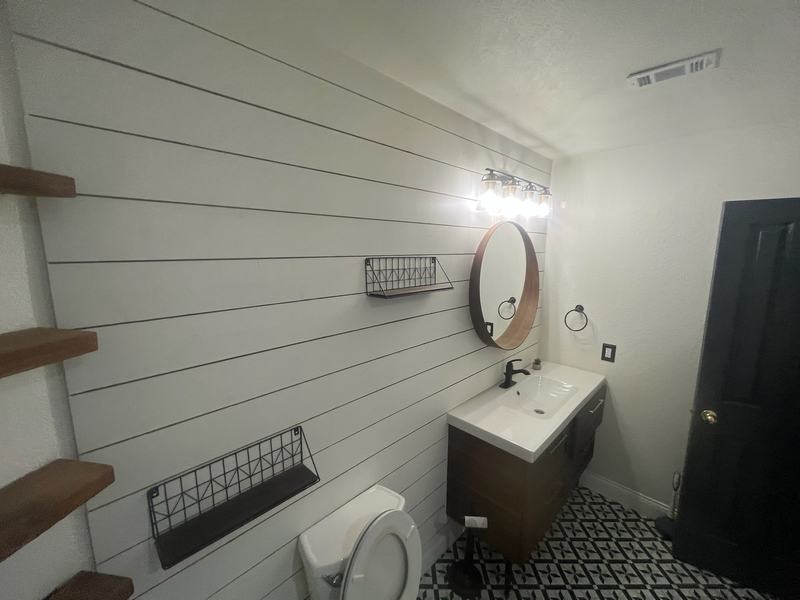 Bathroom with white shiplap walls, floating wood shelves, a round mirror, a vanity and a patterned tile floor