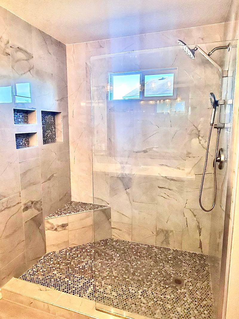 Walk-in shower with large-format marble-look tile, recessed niches, a tiled bench and a pebble floor behind a glass panel
