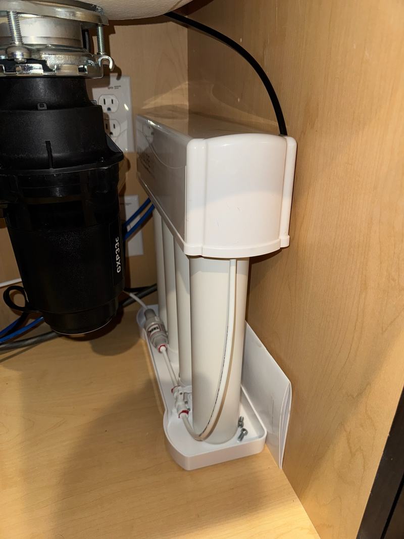 Under-sink reverse-osmosis filtration unit on a cabinet floor with its tubing run, next to a food-waste disposer