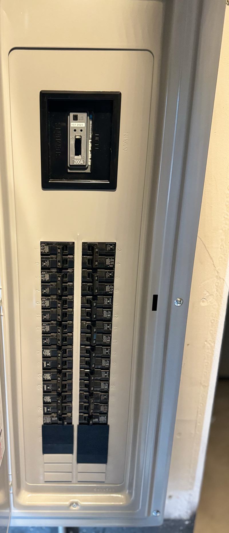 Gray 200-amp breaker panel with its door open, the main breaker at the top and two columns of branch breakers behind the installed cover