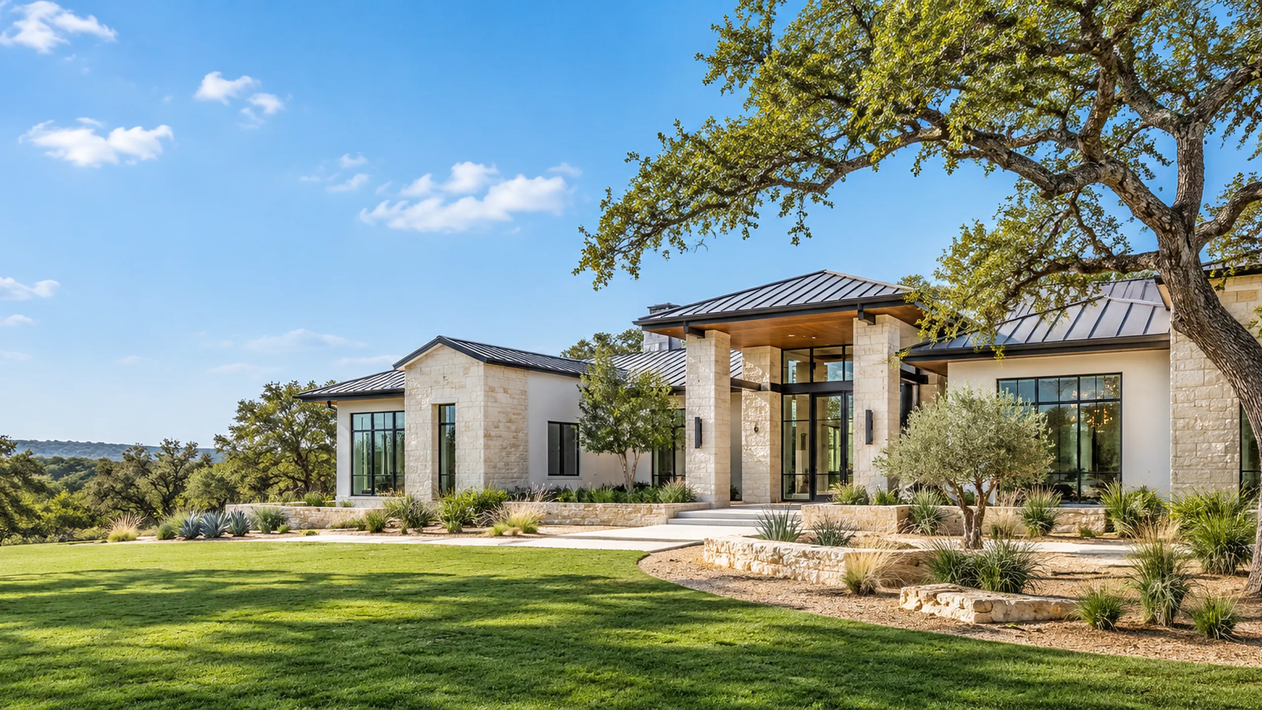 Single-story Hill Country custom home with metal roof in bright daylight, San Antonio TX Single-story cream limestone and white stucco custom home with dark standing-seam metal roofs, tall glass doors, a green lawn, gravel and limestone landscape beds and a large live oak under a bright blue sky
