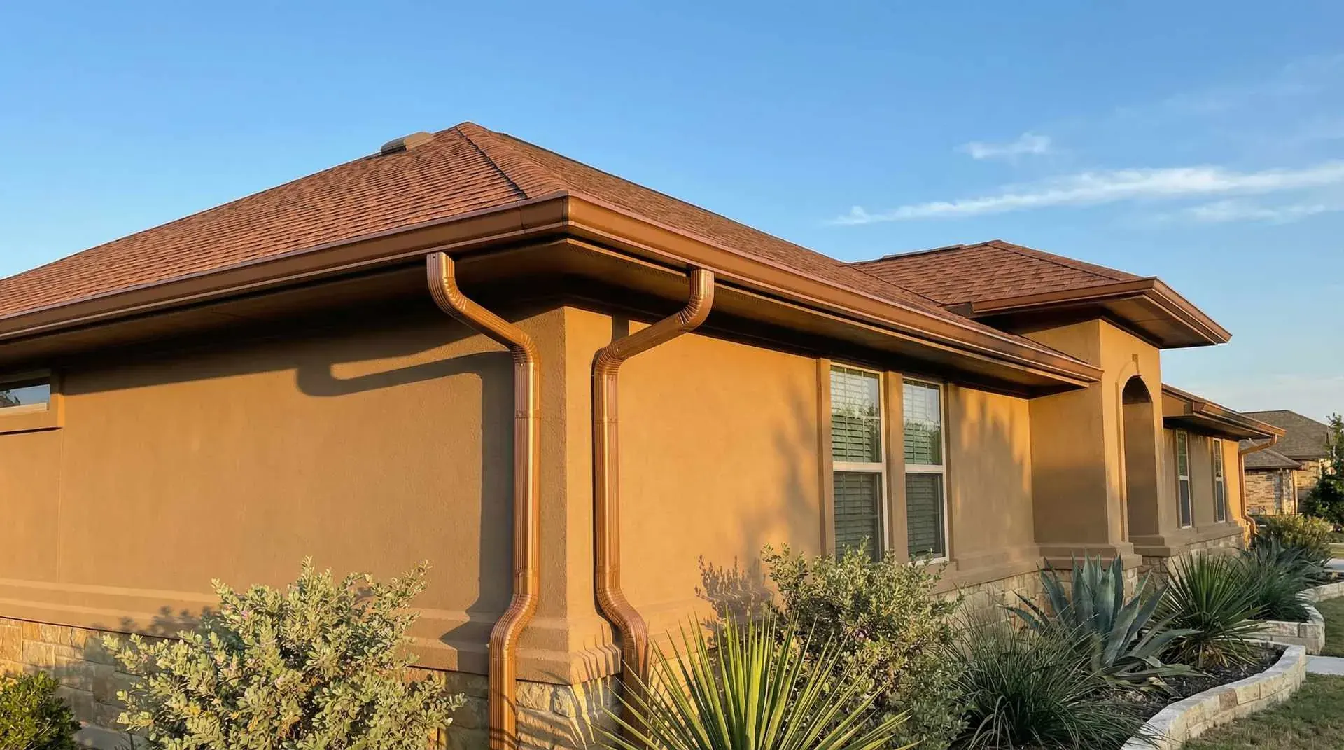 Bronze-finish seamless gutters and two downspouts along the eave of a single-story stucco home.