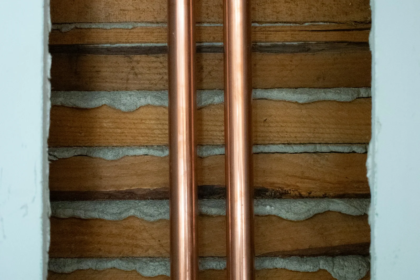 Two vertical copper water pipes exposed in an opened plaster and lath wall.