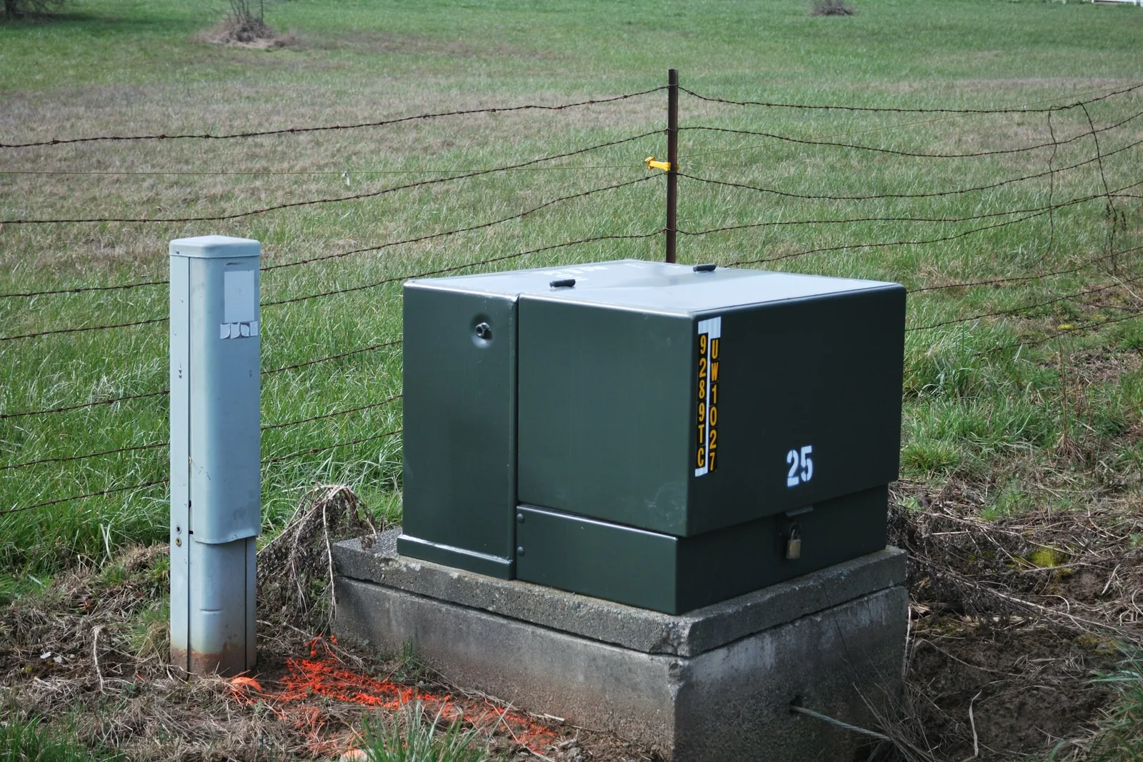 Green pad-mounted utility transformer on a concrete base beside a fenced grass field.